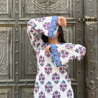 A woman wearing a white long-sleeve tunic dress with pink and blue floral embroidery and blue contrast cuffs, posing in front of a large, weathered wooden door.