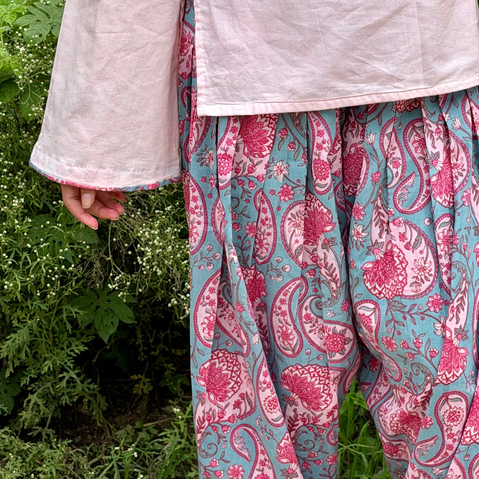A person outdoors wearing a light pink top and loose, patterned pants with a pink paisley print on a teal-blue background, standing in front of green foliage.