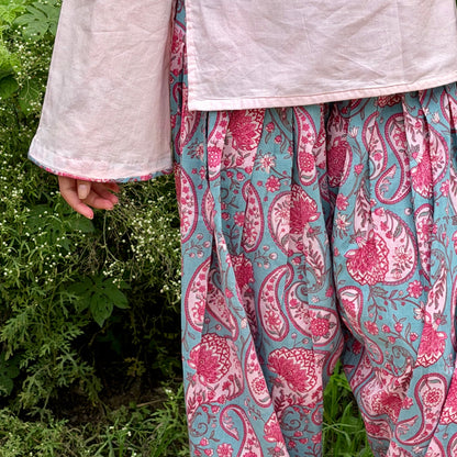 A person outdoors wearing a light pink top and loose, patterned pants with a pink paisley print on a teal-blue background, standing in front of green foliage.