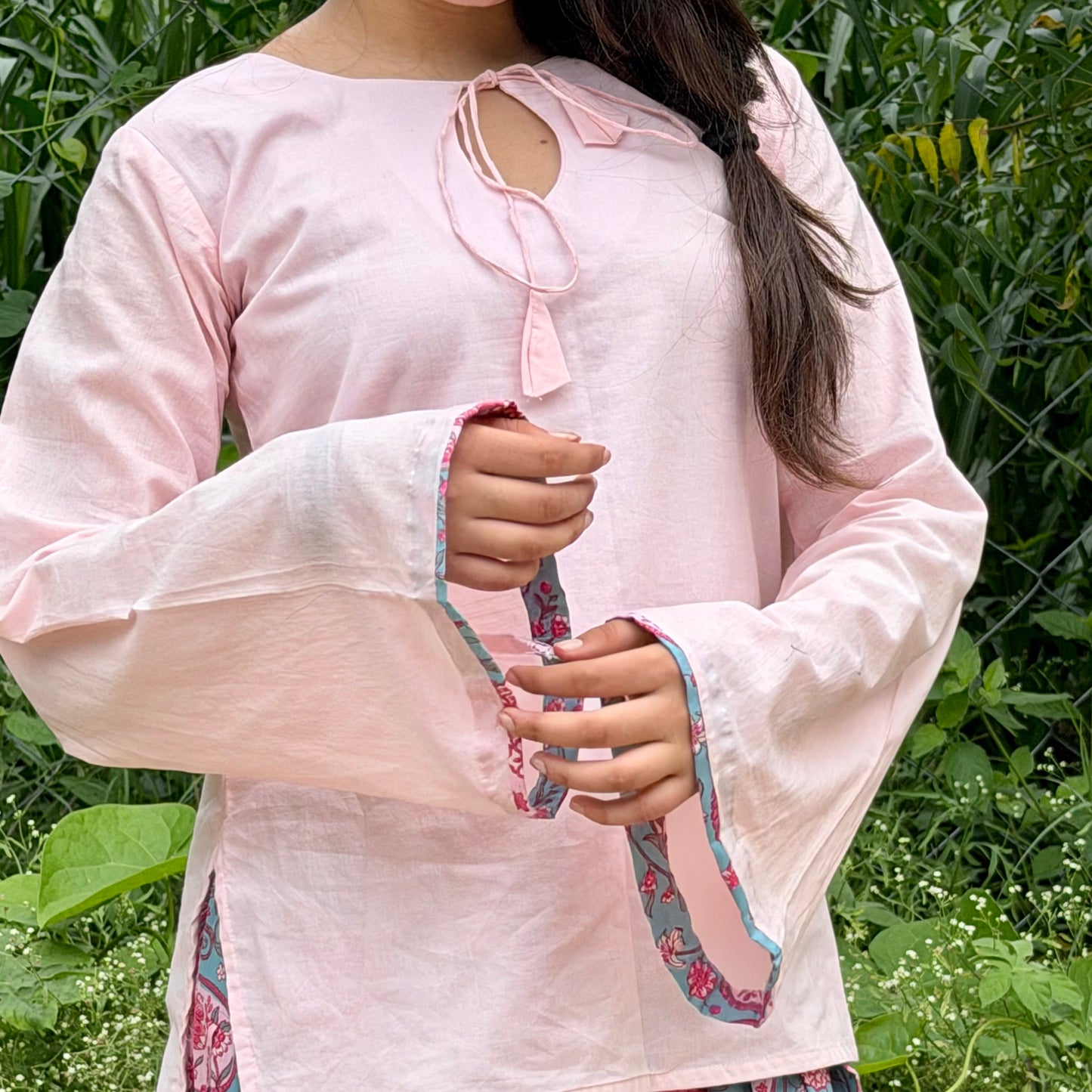 A woman wearing a light pink long-sleeve top with a keyhole neckline and a matching pink paisley skirt, standing outdoors in front of green foliage.