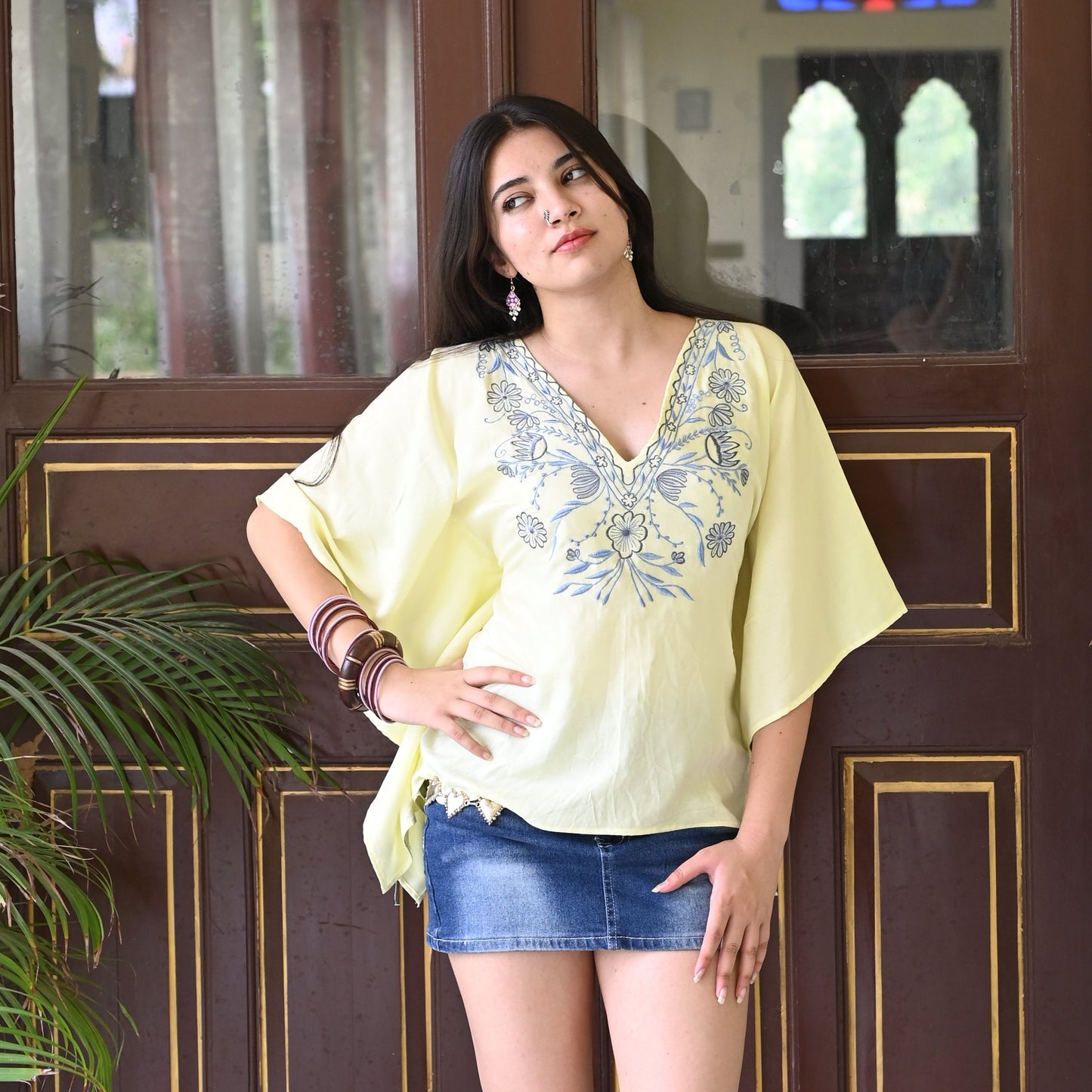 A woman stands in front of a large dark brown wooden door with gold trim, wearing a pale yellow embroidered tunic top, denim shorts, and black knee-high boots. The setting has a warm cream wall, hanging brass lamps, and potted plants.