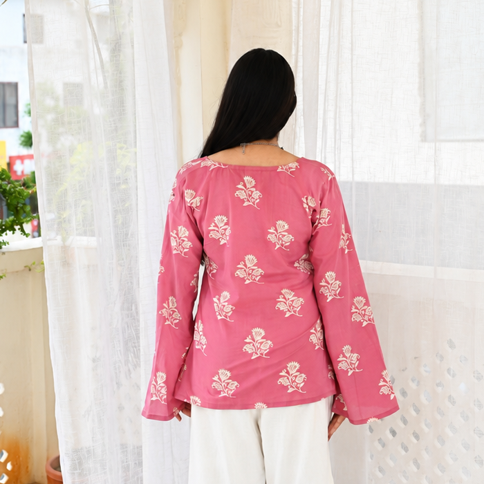 A woman viewed from behind wearing a pink long-sleeve top with a white floral pattern and white wide-leg pants, standing on a balcony with sheer white curtains.