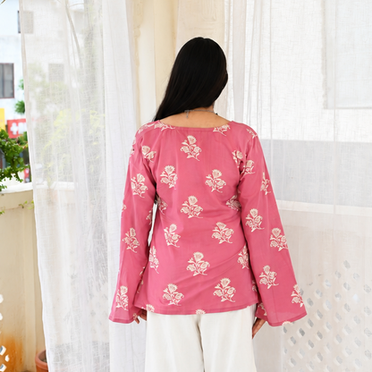 A woman viewed from behind wearing a pink long-sleeve top with a white floral pattern and white wide-leg pants, standing on a balcony with sheer white curtains.
