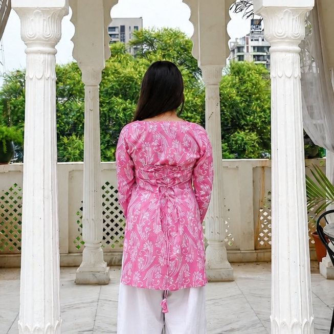 A woman viewed from behind wearing a pink patterned kurta and white trousers, standing inside a white arched pavilion with columns and looking out toward green trees.