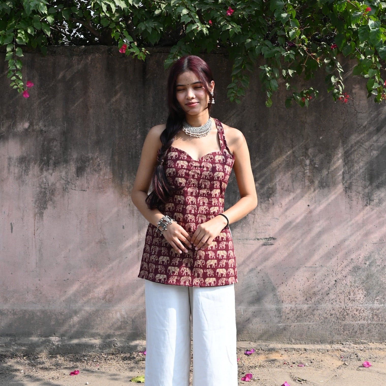 Woman in a patterned top and white pants standing under a tree with pink flowers.