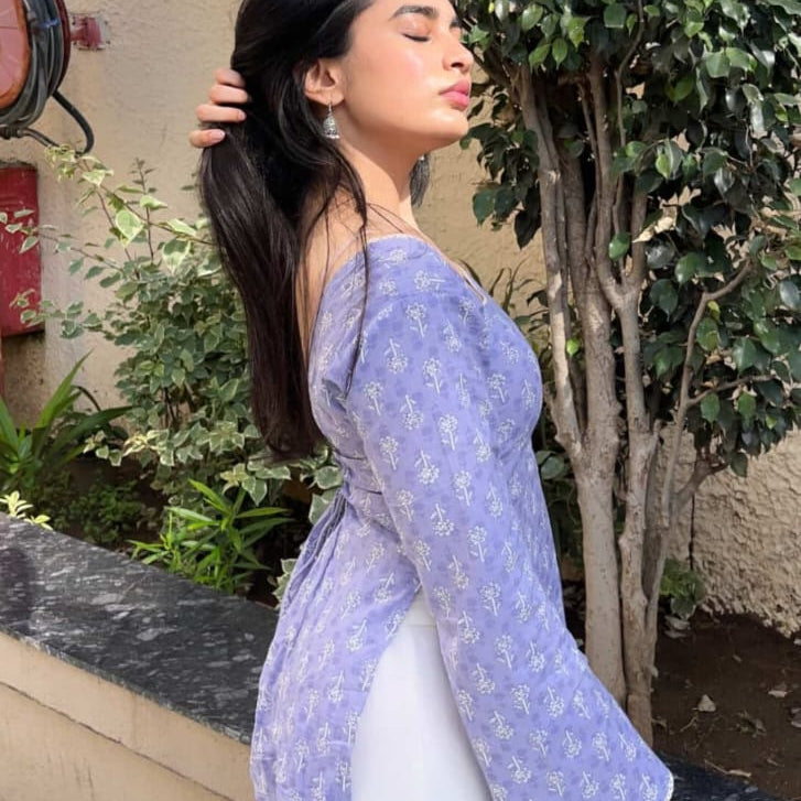 Woman in a blue top and white pants standing outdoors with trees and a building in the background.